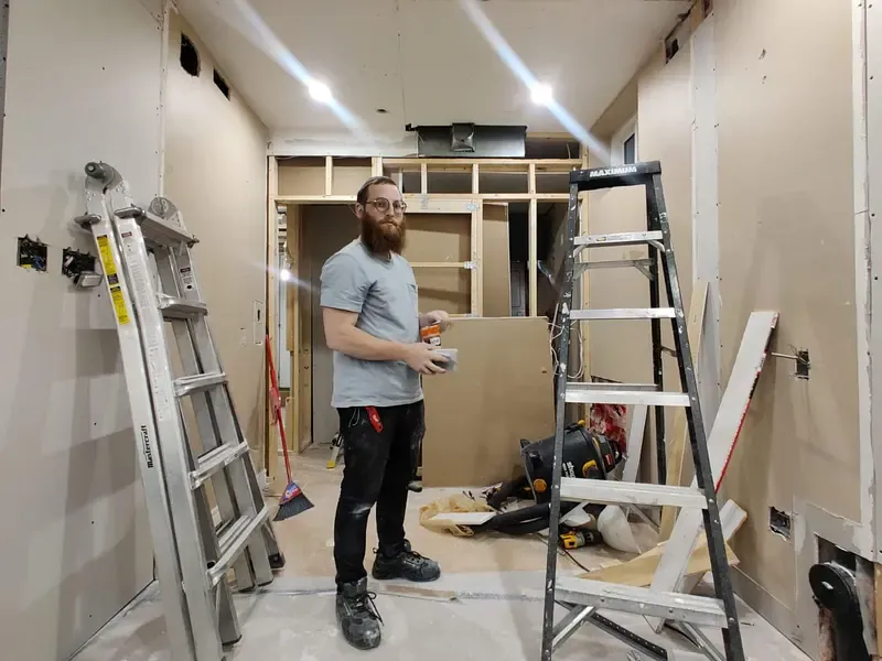 An electrician on a renovation site, with open framing, ladders, and a panel opening